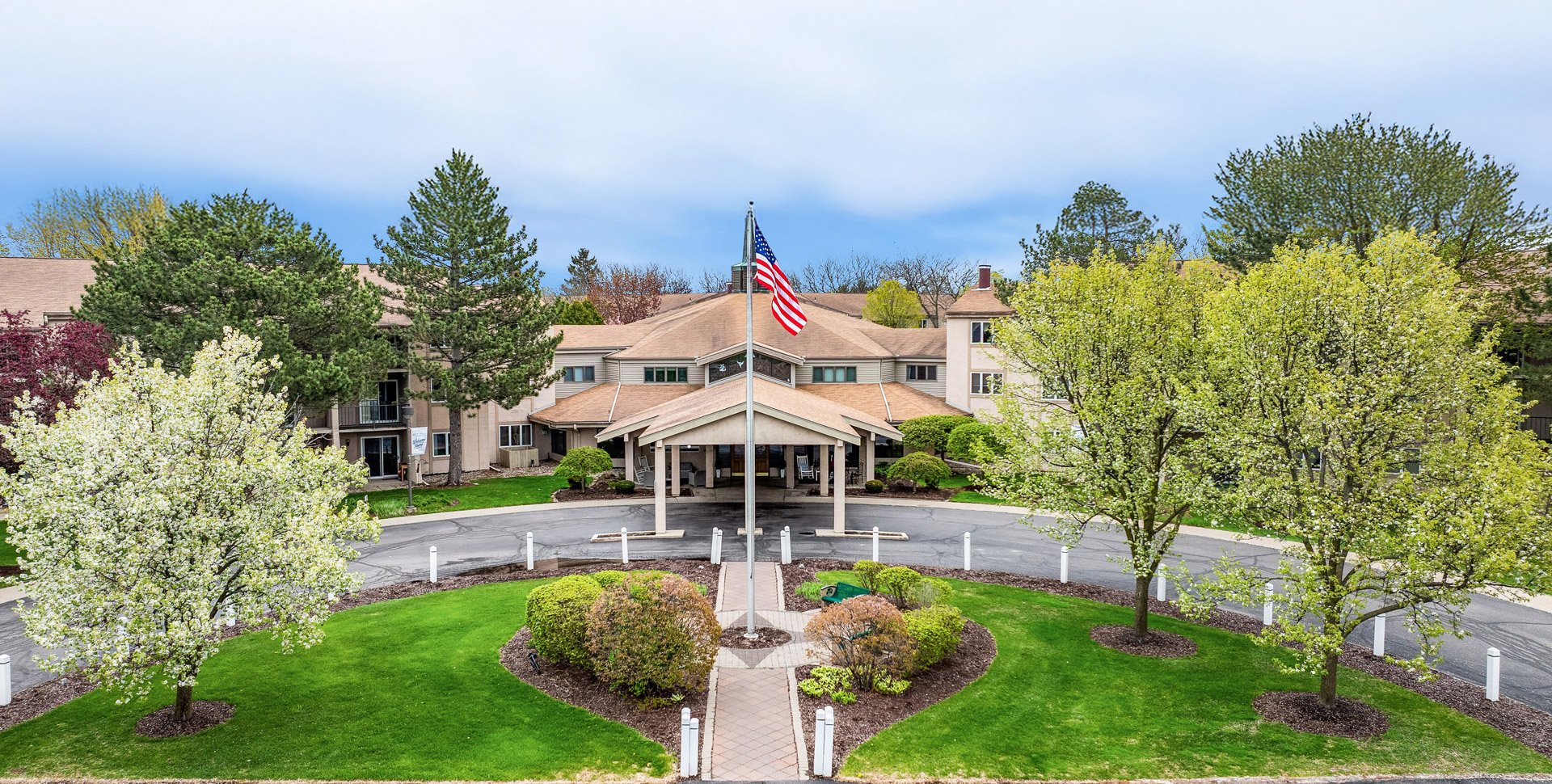 A large building with a circular driveway, landscaped greenery, and an American flag at the entrance. Trees and bushes surround the driveway under a partly cloudy sky.